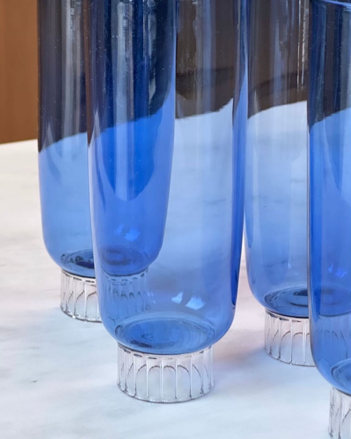 The image offers a detailed view of colored champagne glasses, highlighting their exquisite cobalt blue colored glass. The lower section of each flute displays a beautiful stemmed bottom, intricately designed with a ribbed texture that captures and bends the light, adding a touch of elegance and visual interest. The flutes transition from this ornate base into a striking cobalt blue color, which gives these colored champagne glasses a vibrant and luxurious appearance.