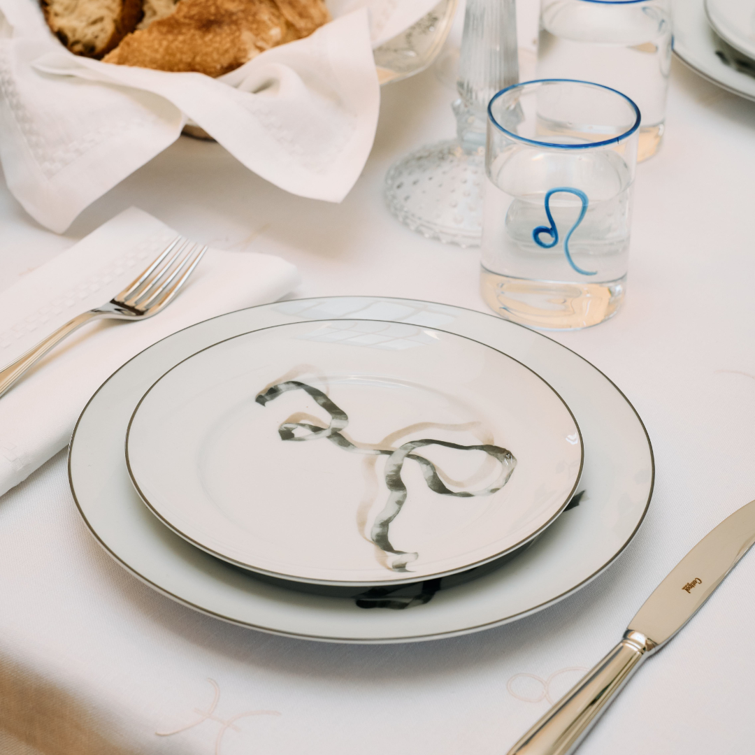 salad plate with a black ribbon placed atop a matching dinner plate. To the left, a neatly folded white napkin lies under a silver fork, and to the right, a silver knife with a refined design. In the background, transparent hand-blown glass with cobalt blue rim and Leo astrology sign. The overall setting exudes a sense of quiet elegance.