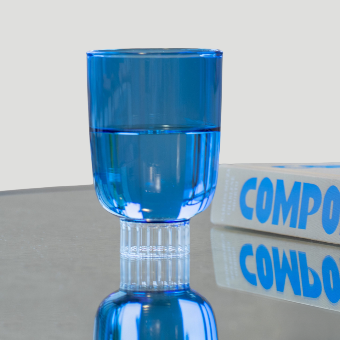 A vibrant cobalt blue drinking glass from our colored glassware collection is elegantly displayed, filled halfway with water, on a mirror coffee table. Made from 100% borosilicate glass, it is lightweight yet durable. The perfect gift for everyone in your life