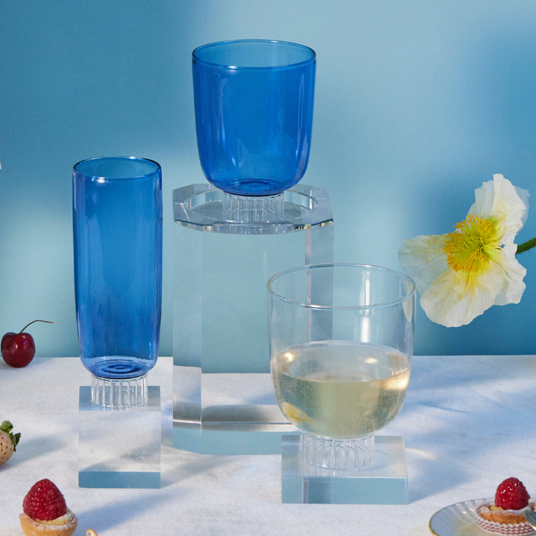 A Beautifully composed table setting that radiates a romantic vibe. At the heart of the setting is an assortment of glassware, including cobalt blue champagne flutes, clear cocktail glasses,  as well as elegant stemless wine glasses, with clear textured stems, all lending a modern and sophisticated touch. These glasses are dishwasher safe and stackable. The table also features a selection of beautiful flowers, fresh fruits and gourmet pastries, which infuse the scene with a sense of abundance and delight.