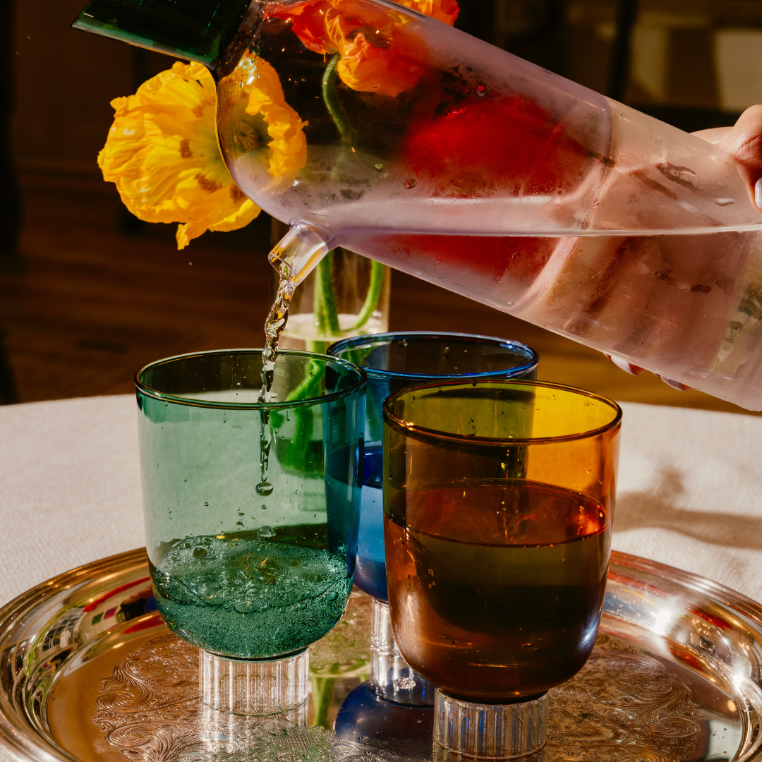 Discover Sprezz's premium colored glassware, water is being poured from a color blocked lilac water pitcher into a stemless teal colored glass. The image features a luxurious silver tray with a set of Sprezz's Romantic vintage-style glasses in rich hues of forest green, cobalt blue, and amber. The romantic ambiance is perfected by a softly lit candle, highlighting the intricate designs and exceptional craftsmanship that define the Sprezz glassware collection.