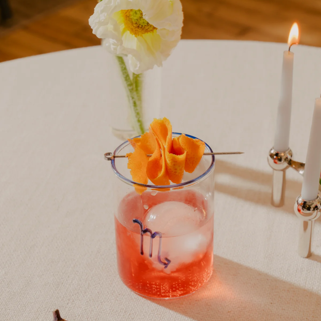 Scorpio zodiac glass with a Negroni cocktail, blue rim, and cobalt symbol, styled with figs, a white poppy, and candlelight for a sophisticated, sensual date night vibe.