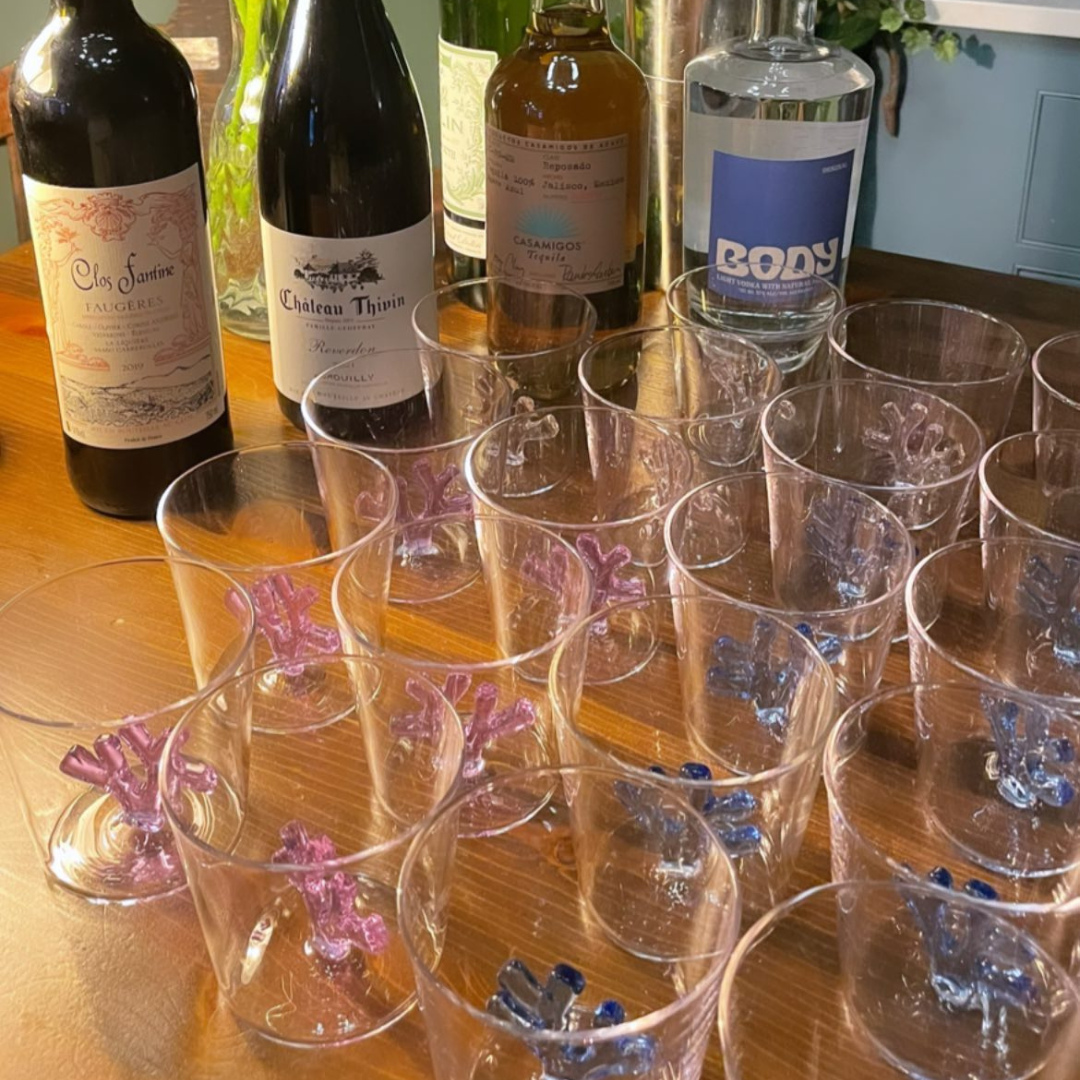 An array of Sprezz unique glassware is artfully displayed on a wooden bar, with coral sculptures in shades of lilac and blue inside of the glass. The scene is set with an eclectic mix of wine and spirits bottles, a bouquet of pink ranunculus flowers in a glass vase,  creating a vibrant and inviting atmosphere for memorable dinner parties