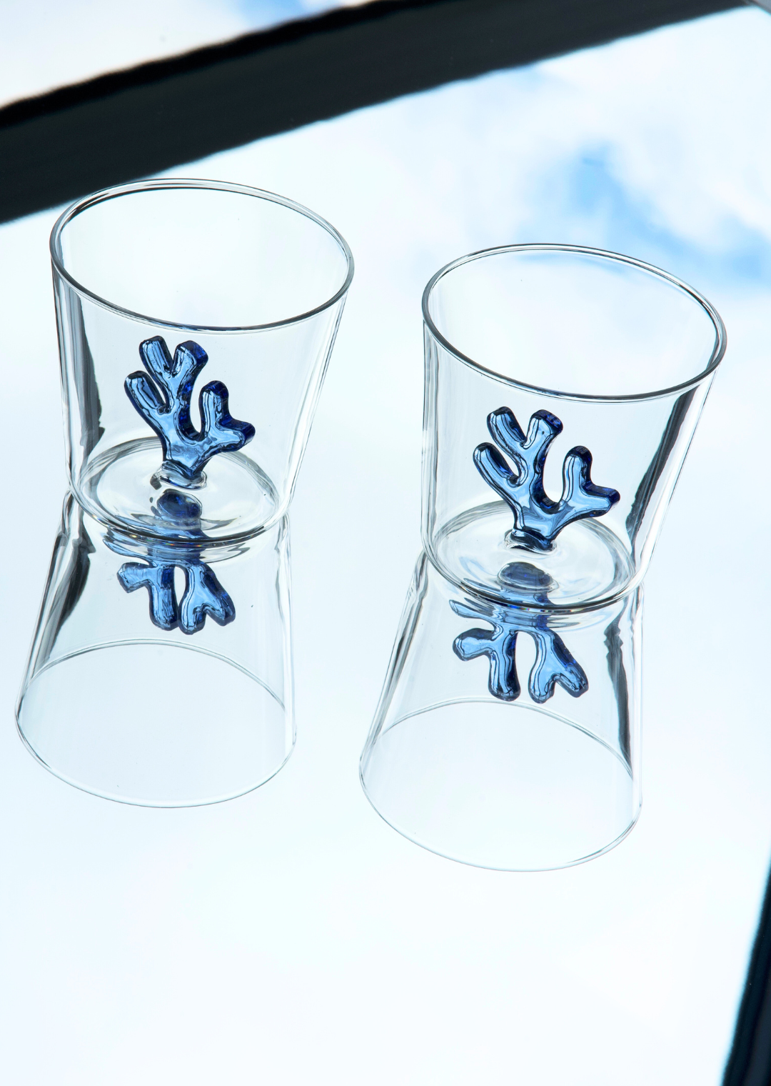 A set-of-two water tumblers on a mirrored table. The Sprezz all-purpose water tumblers features an blue coral morif on the inside