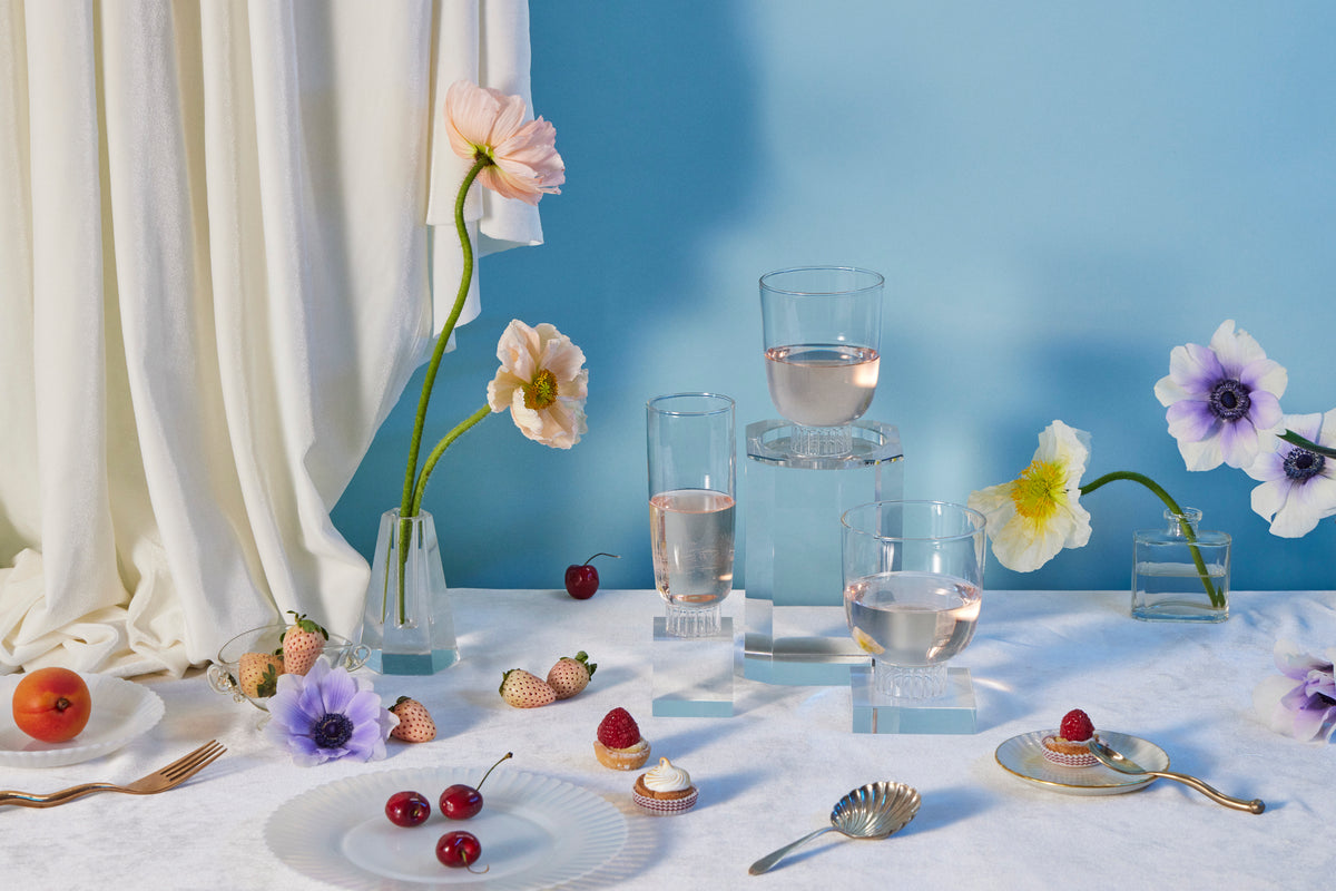 Elegant table setting featuring Sprezz's clear cocktail glasses, stemless wine glasses, and champagne glasses. Hand-blown from high-quality, dishwasher-safe,  borosilicate glass, these lead-free non-toxic glasses are complemented by an arrangement of fresh flowers, strawberries, and pastries. 