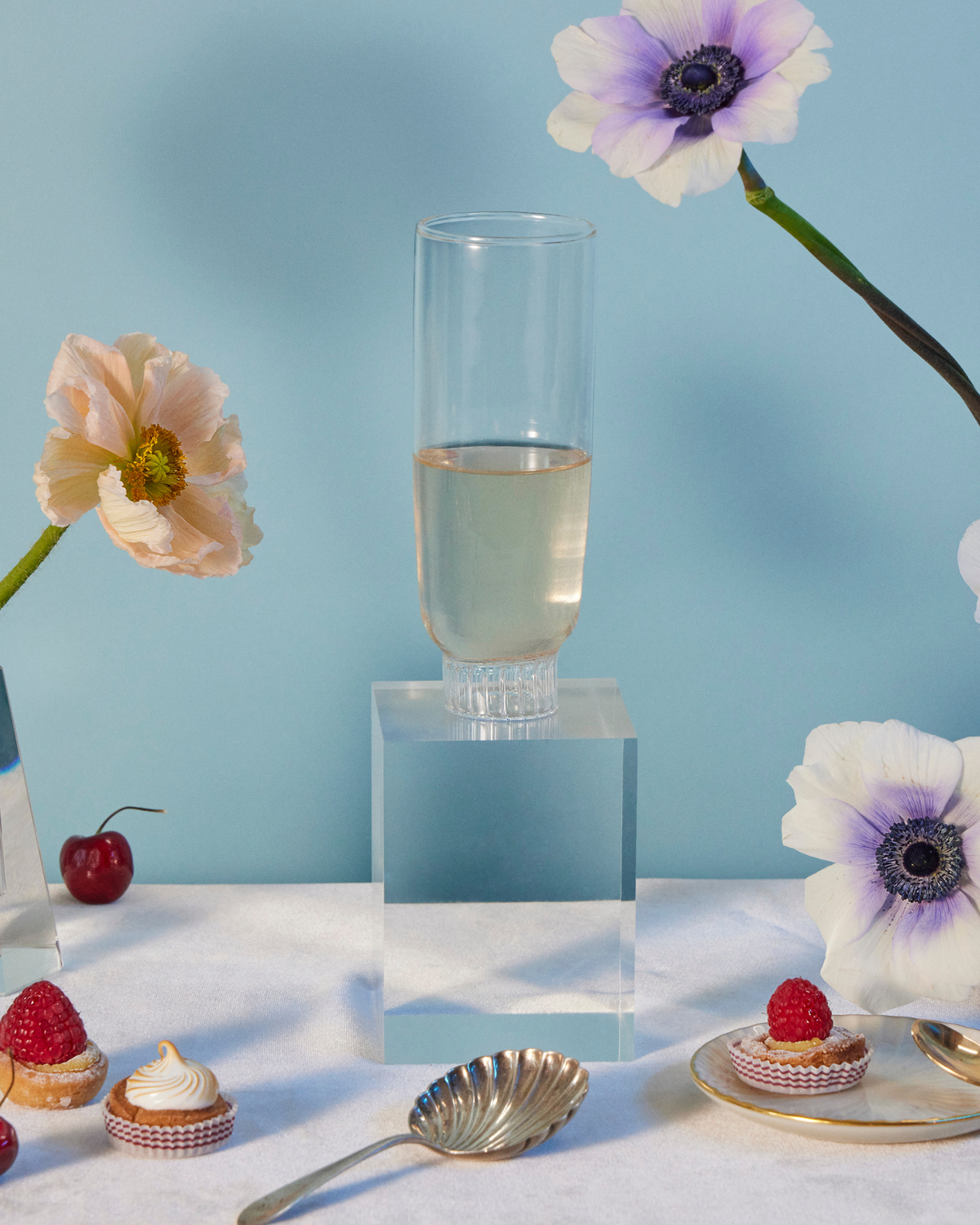 Sprezz's borosilicate glass champagne flute, amid an elegant table setting with pale pink poppies and purple-centered anemones in glass vases, fresh strawberries, cherries, and delicate pastries on a gold-accented white tablecloth with a serene blue backdrop.These durable, thermal shock-resistant glasses embody modern elegance, perfect for luxury dining and as the most thoughtful gifts for friends and family.