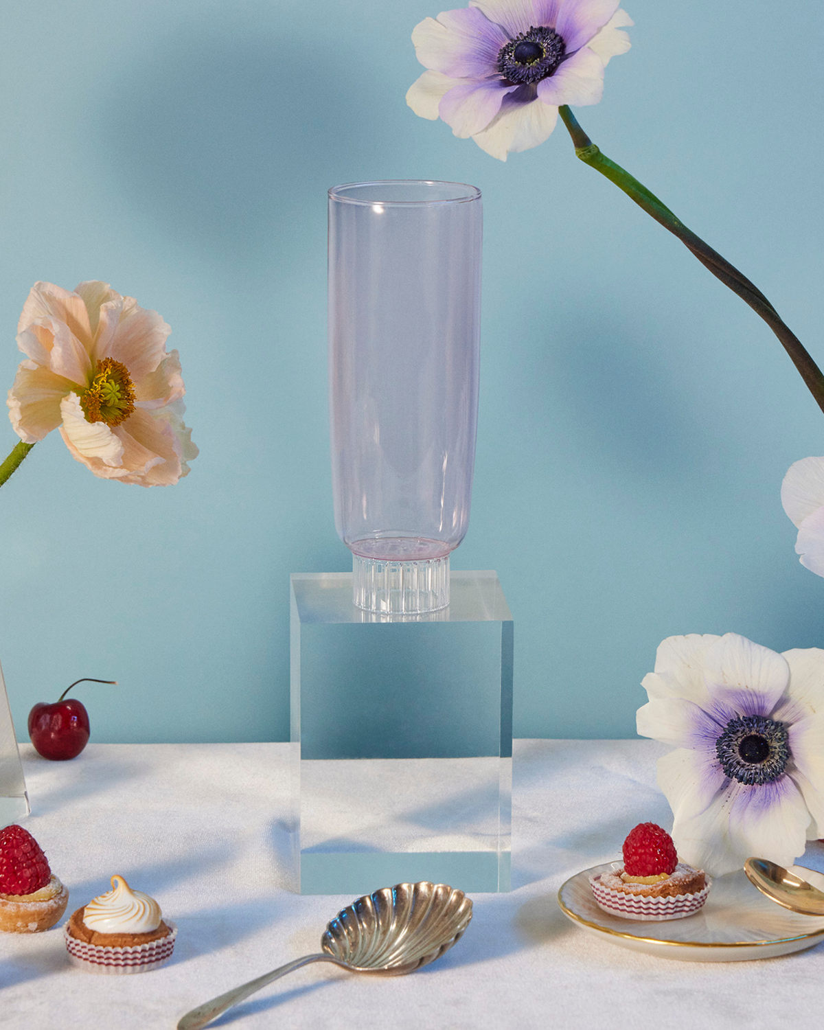 Sprezz's hand-blown colored champagne glass, crafted from dishwasher-safe, high-quality borosilicate glass, on an elegant table setting with pale pink poppies and purple-centered anemones in glass vases, fresh strawberries, cherries, and delicate pastries. These blush pink flute exudes effortless elegance, its perfect for girl dinners and makes the most thoughtful gifts for friends and family.