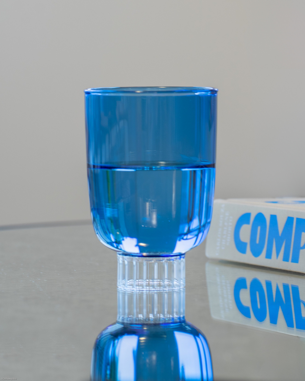 A vibrant cobalt blue drinking glass from our colored glassware collection is elegantly displayed, filled halfway with water, on a mirror coffee table. Made from 100% borosilicate glass, it is lightweight yet durable. The perfect gift for everyone in your life 