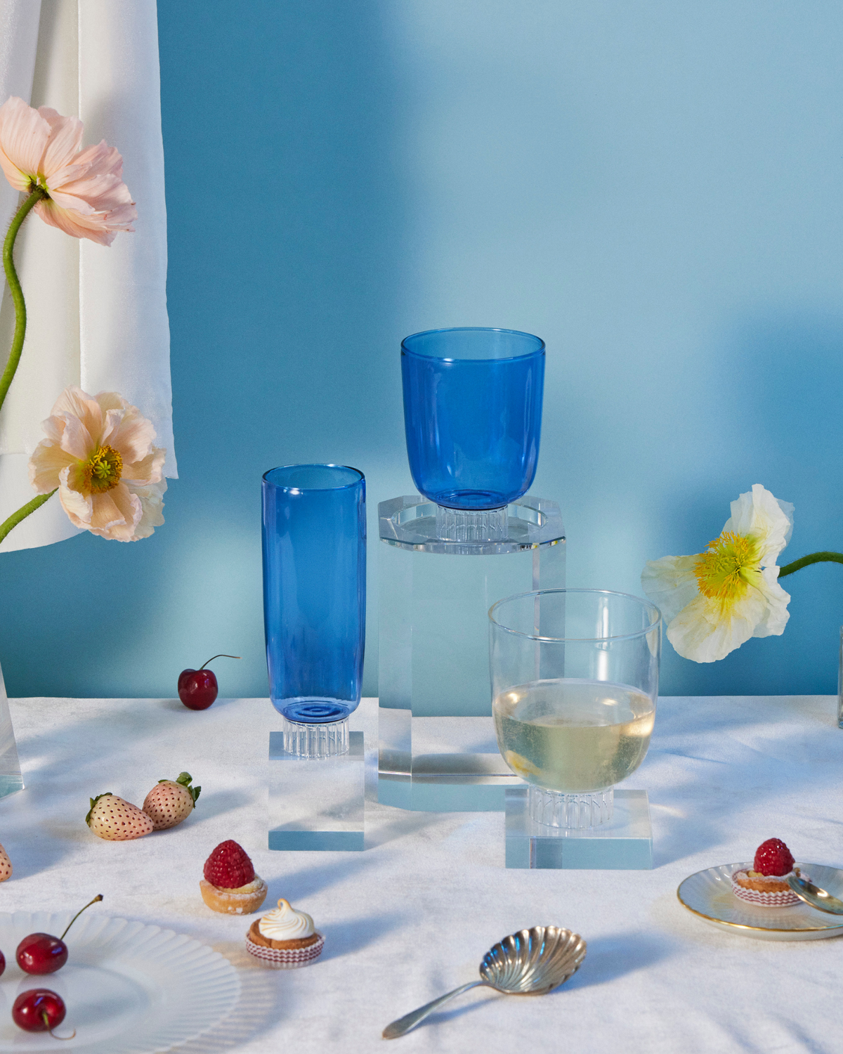 A Beautifully composed table setting that radiates a romantic vibe. At the heart of the setting is an assortment of glassware, including cobalt blue champagne flutes, clear cocktail glasses,  as well as elegant stemless wine glasses, with clear textured stems, all lending a modern and sophisticated touch. These glasses are dishwasher safe and stackable. The table also features a selection of beautiful flowers, fresh fruits and gourmet pastries, which infuse the scene with a sense of abundance and delight.