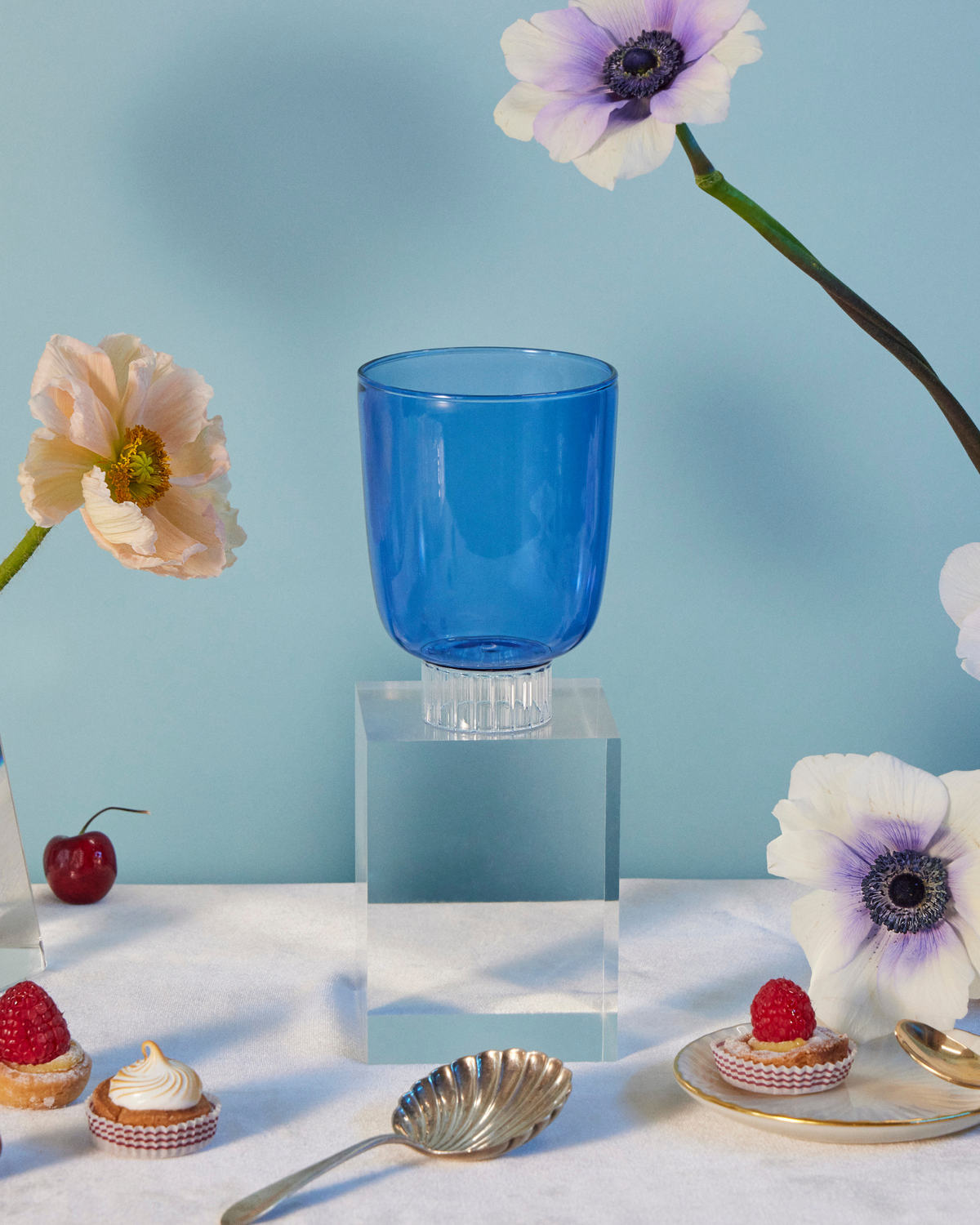 Set-of-four cobalt blue colored stemless wine glasses. A colored glass on a table with poppies and anemones flowers, cherries, raspberry tarts, rose strawberries, and grapes. 