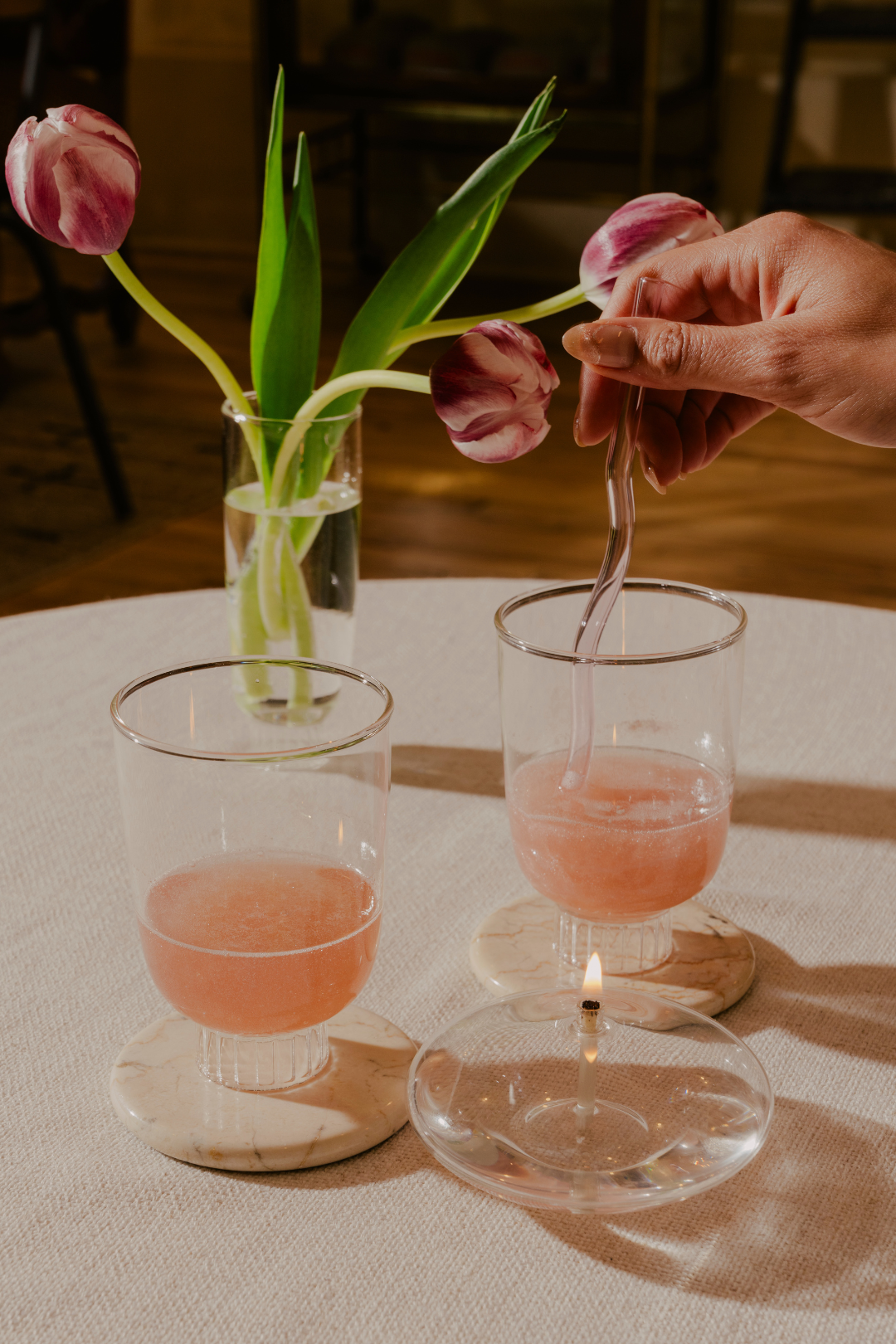 Set of two stemless wine glasses with rose sprinkled with glitter being gently stirred with a blush pink glass straw. The stemless wine glass sits on a marble coaster, its simplicity and elegance highlighted by the warm glow of the candle. The clear champagne glasses has been used as bud vase with slender tulips. This scene suggests a relaxed yet sophisticated setting, possibly a romantic dinner or a cute dinner with the girlies.