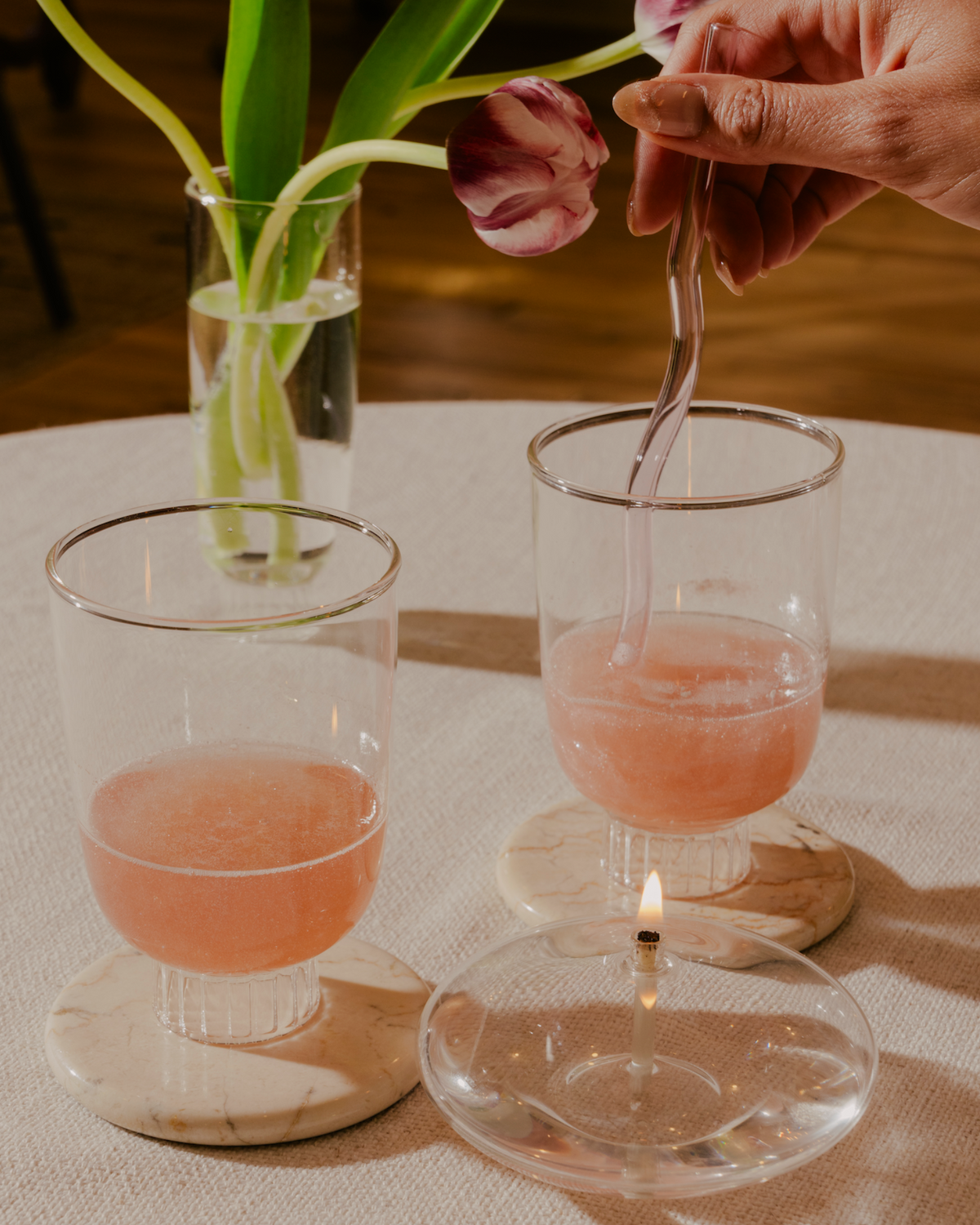 Set of two stemless wine glasses with rose sprinkled with glitter. The stemless wine glass sits on a marble coaster. Made of 100% borosilicate glass it is lightweight yet durable and dishwasher safe. The clear champagne glasses has been used as bud vase with slender tulips. This scene suggests a relaxed yet sophisticated setting, possibly a romantic dinner or a cute dinner with the girlies.