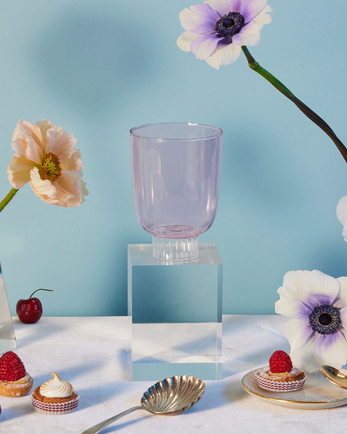 Sprezz colorful stemware. Blush pink colored water glass on a table with poppies and anemones flowers, cherries, raspberry tarts, rose strawberries, and grapes. Made from 100% borosilicate