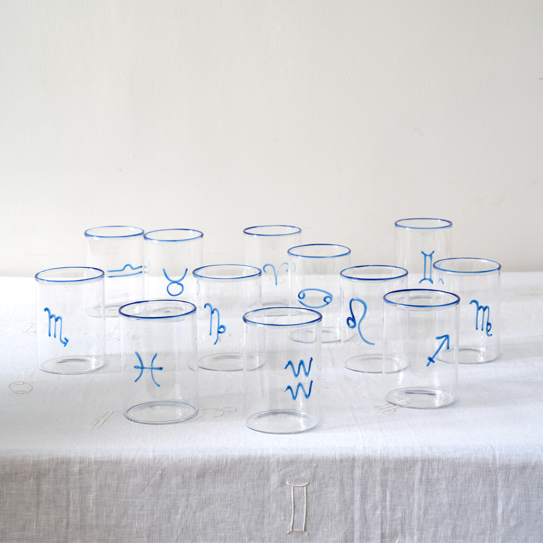A collection of clear rocks glasses, each adorned with a different blue astrological symbol and cobalt blue rim. The glasses are arranged in rows on a linen-textured tablecloth, exhibiting the 12 zodiac signs —  Aquarius, Pisces, Aries, Cancer, Taurus, Virgo, Capricorn, Scorpio, Sagittarius, Libra, Leo, Gemini. Though they feel modern, they’re elegant enough to work with just about any tableware from just about any era. 