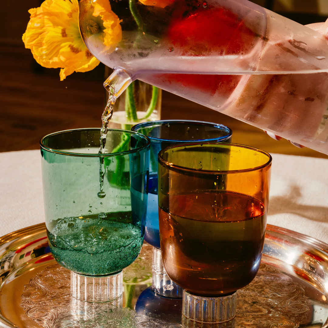 Discover Sprezz's premium colored glassware, water is being poured from a color blocked lilac water pitcher into a stemless teal colored glass. The image features a luxurious silver tray with a set of Sprezz's Romantic vintage-style glasses in rich hues of forest green, cobalt blue, and amber. 