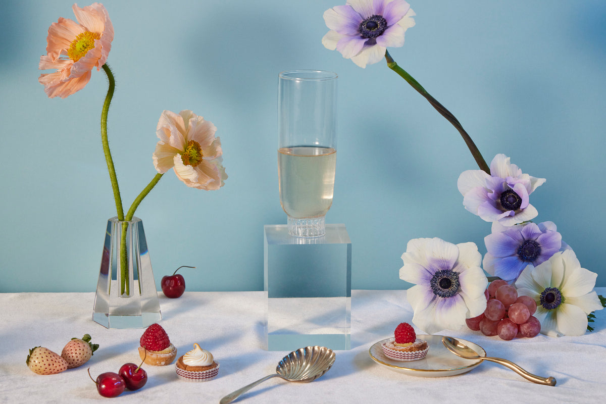 Sprezz's borosilicate glass champagne flute, amid an elegant table setting with pale pink poppies and purple-centered anemones in glass vases, fresh strawberries, cherries, and delicate pastries on a gold-accented white tablecloth with a serene blue backdrop.These durable, thermal shock-resistant glasses embody modern elegance, perfect for luxury dining and as the most thoughtful gifts for friends and family.