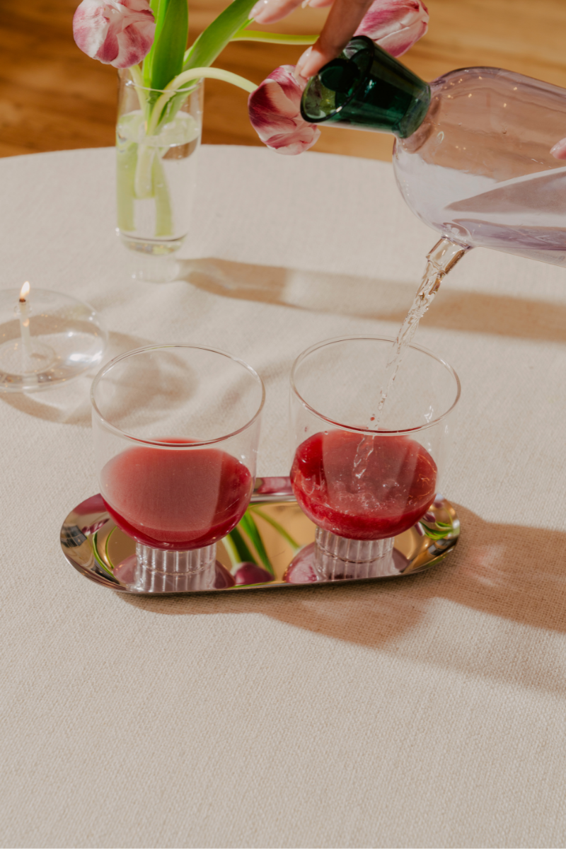 Two lead-free, clear stemless wine glasses containing vibrant red beverages. These hand-blown borosilicate cocktail glasses are set on a reflective tray alongside a vase of pink tulips and a lit tealight candle, on a natural linen tablecloth, creating a warm, inviting ambiance for your next date night.