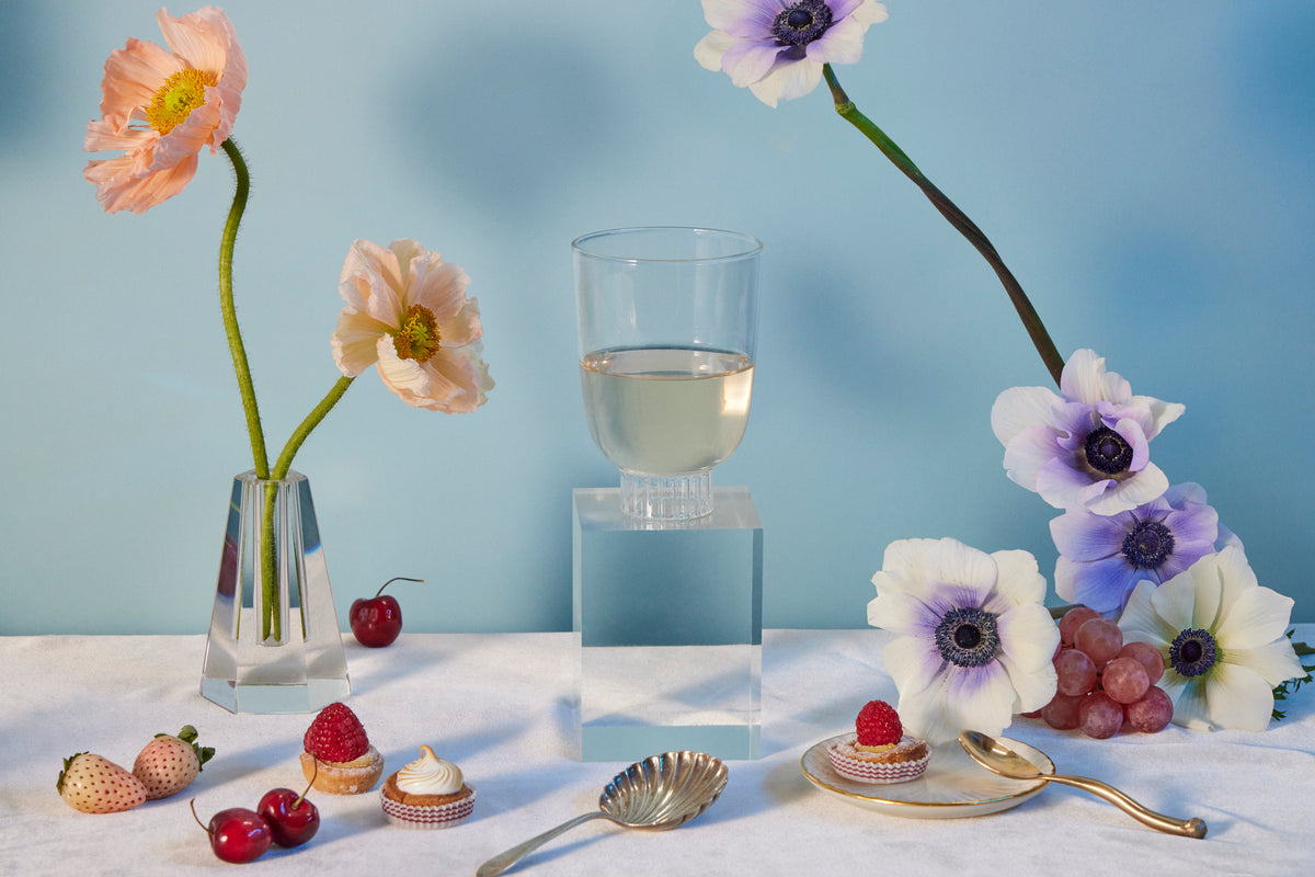 Sprezz romantic clear wine glass on a table with poppies and anemones flowers, cherries, raspberry tarts, rose strawberries, and grapes. 