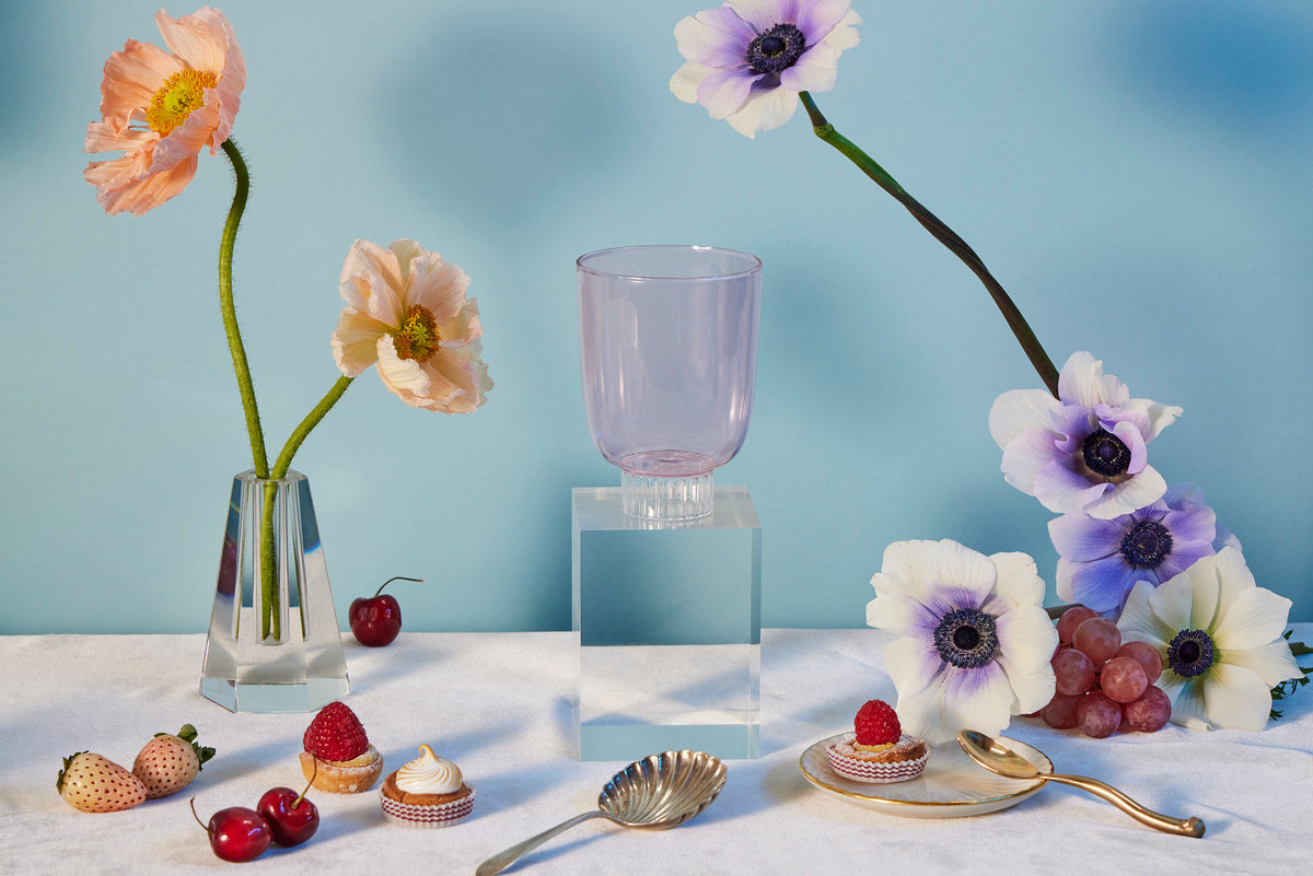 Sprezz colorful stemware. Blush pink colored water glass on a table with poppies and anemones flowers, cherries, raspberry tarts, rose strawberries, and grapes. 