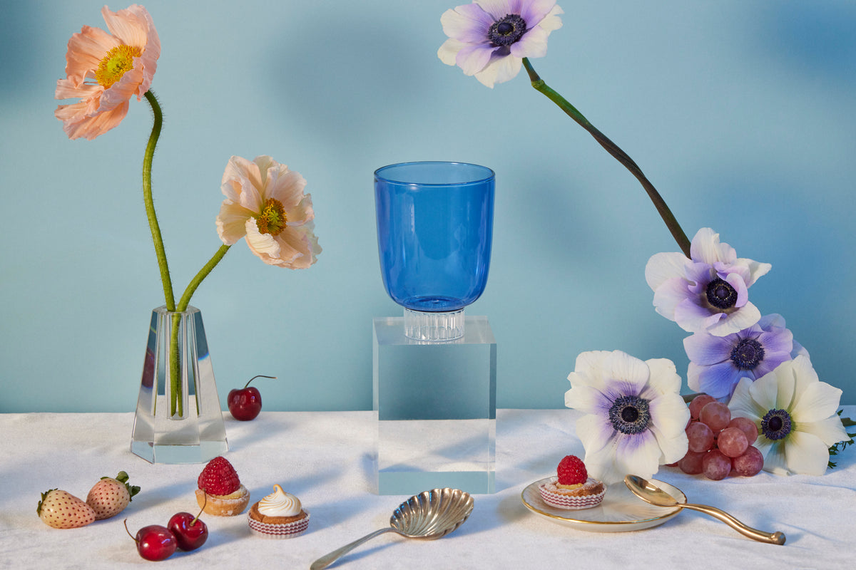 Set-of-four cobalt blue colored water glass on a table with poppies and anemones flowers, cherries, raspberry tarts, rose strawberries, and grapes. 
