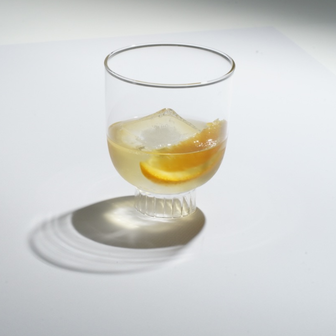 This image captures the contemporary, minimalist allure of a Sprezz glass with a modernist design, featuring geometric lines and an original cylindrical stem. Ideal for red wine, sipping tequilla, whiskey, the glass is made from high-quality, lead-free borosilicate, offering exceptional transparency and brilliance. The drink, is accented with a large ice cube and orange slice, emphasizes the glass's elegant profile