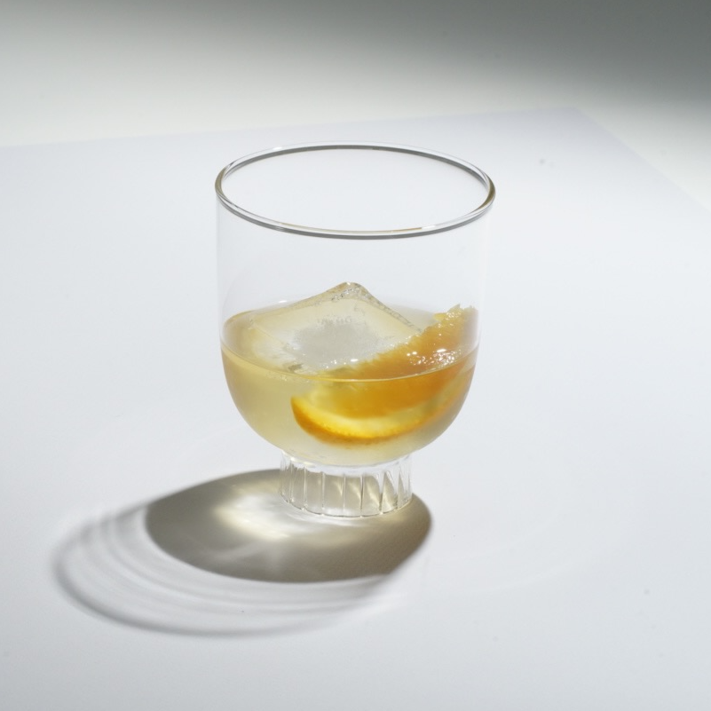 This image captures the contemporary, minimalist allure of a Sprezz glass with a modernist design, featuring geometric lines and an original cylindrical stem. Ideal for red wine, sipping tequilla, whiskey, the glass is made from high-quality, lead-free borosilicate, offering exceptional transparency and brilliance. The drink, is accented with a large ice cube and orange slice, emphasizes the glass's elegant profile