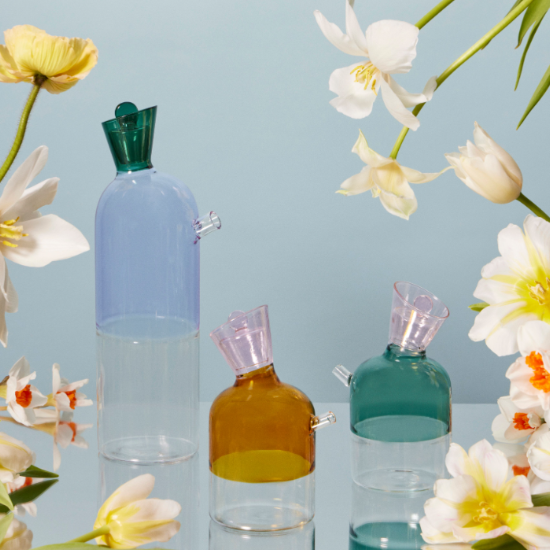 Lilac Carafe and two oil and vinegar bottles in teal and amber borosilicate glass on a mirrored table surrounded by tulips and daffodils. Contemporary, minimalist allure animates the modernist design of these colored glass decanters