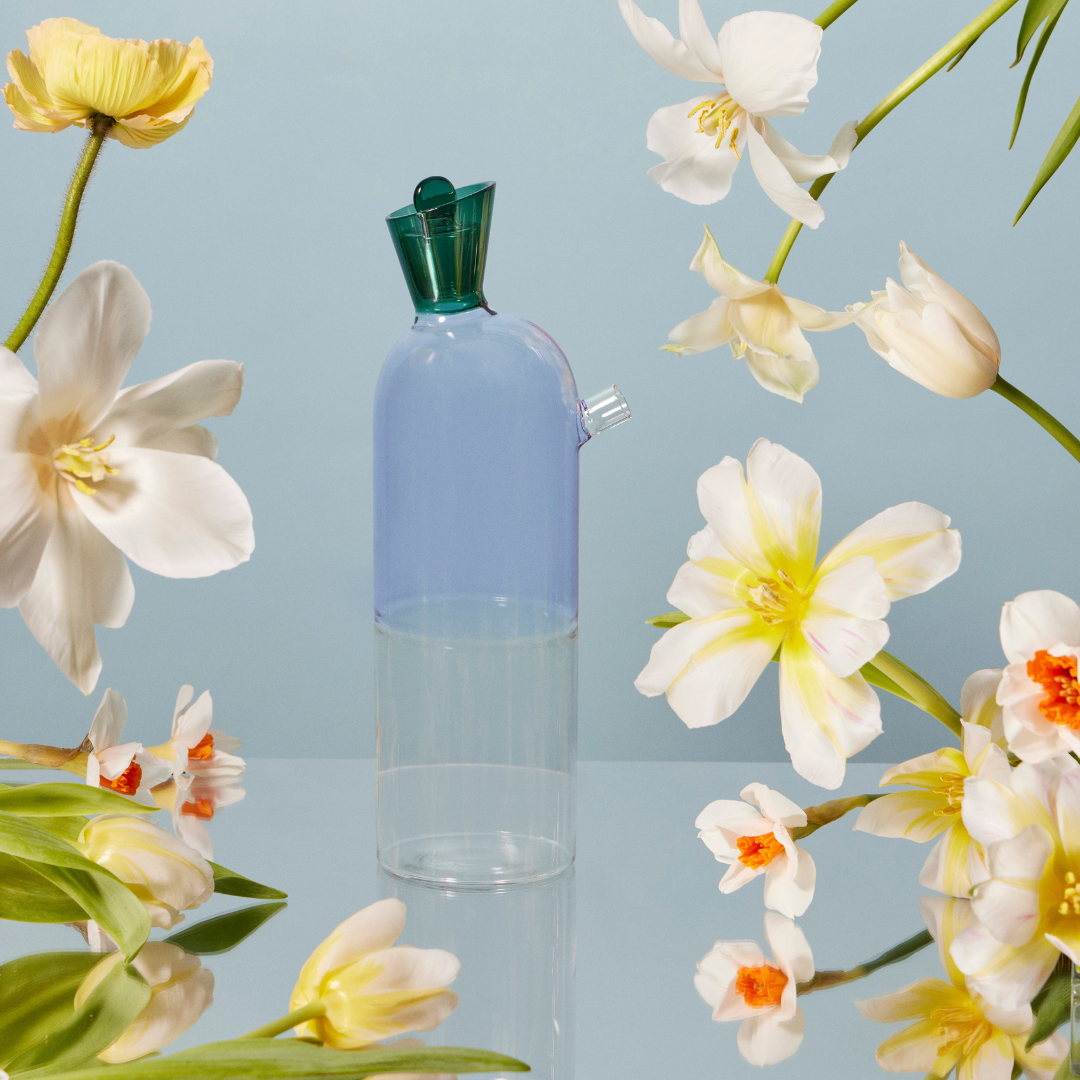 A whimsical lilac carafe and pitcher with a tapered body and anemerald green stopper, on a mirrored table. The decanter is surrounded by beautiful creamy white and soft yellow flowers, adding a touch of whimsical elegance the dinner table. 