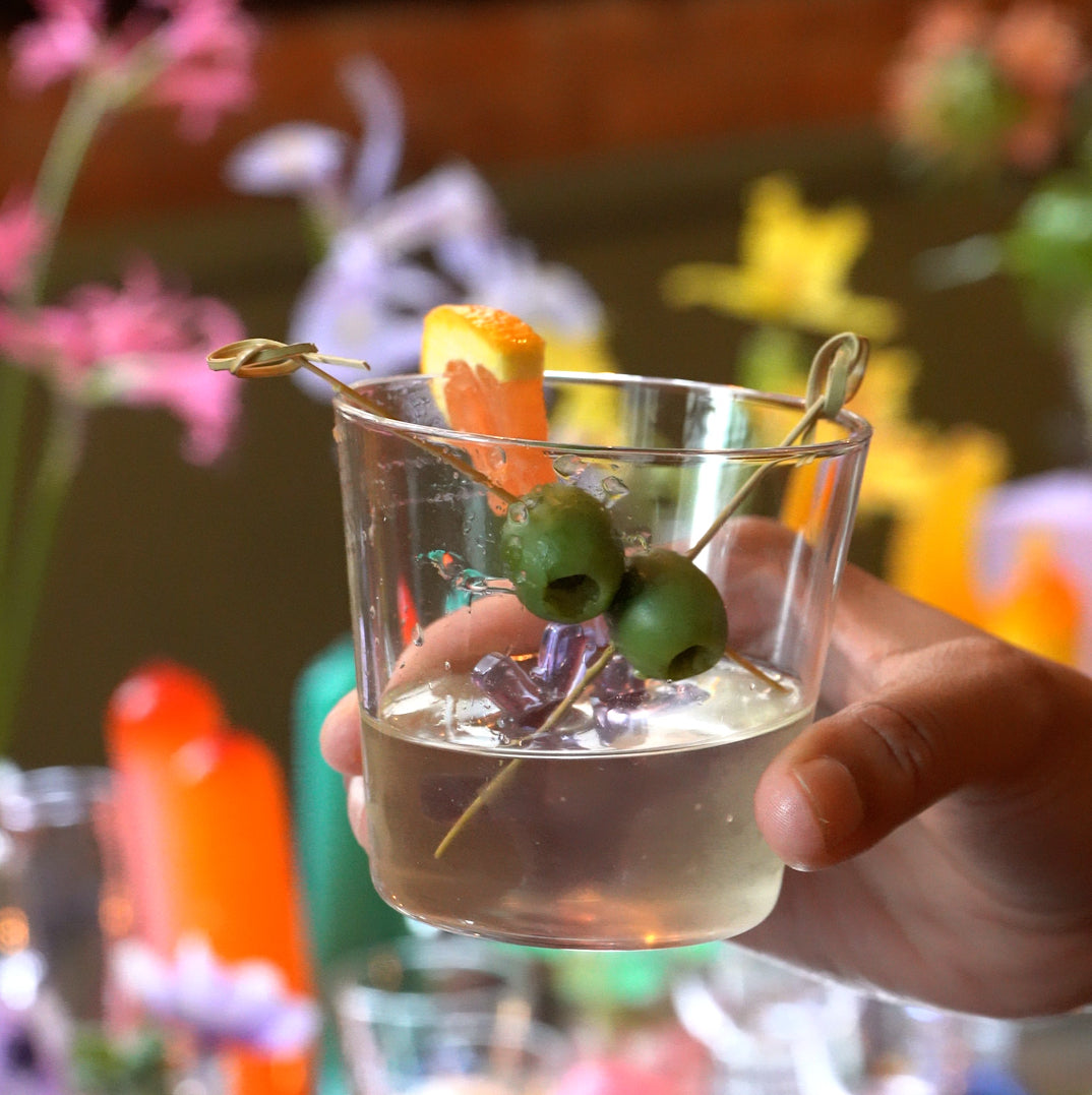 A hand holding a colored martini glass infront of a beautiful dinner table with lots of flowers