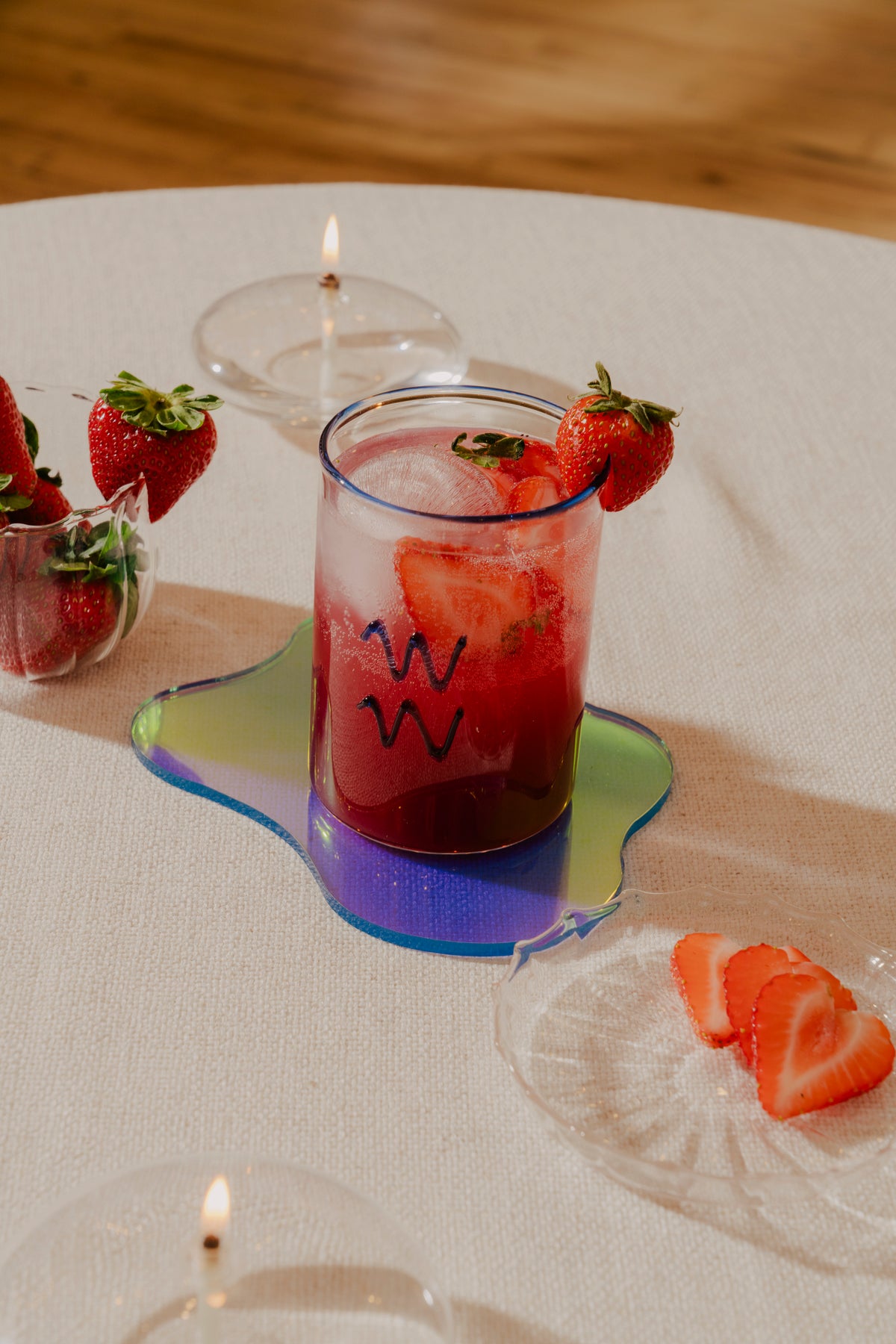 A refreshing  non-alcoholic strawberry spritz in a Sprezz Aquarius Lowball Glass, artistically garnished with a whole strawberry on the rim, sits on a colorful coaster. This vibrant scene is completed with a bowl of fresh strawberries and a lit candle, adding a cozy and inviting ambiance to this elegant table setup.