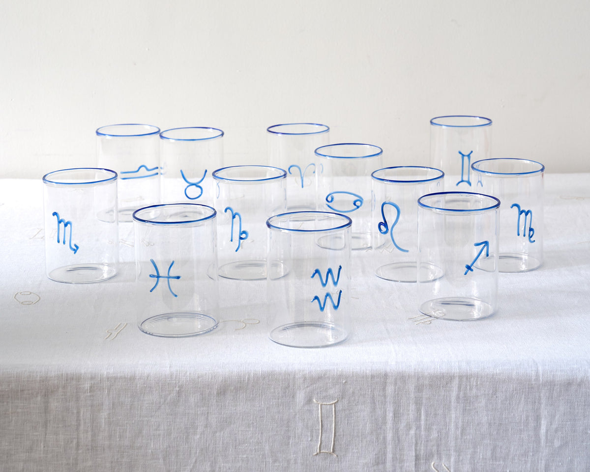 A collection of clear rocks glasses, each adorned with a different blue astrological symbol and cobalt blue rim. The glasses are arranged in rows on a linen-textured tablecloth, exhibiting the 12 zodiac signs —  Aquarius, Pisces, Aries, Cancer, Taurus, Virgo, Capricorn, Scorpio, Sagittarius, Libra, Leo, Gemini. Though they feel modern, they’re elegant enough to work with just about any tableware from just about any era. 