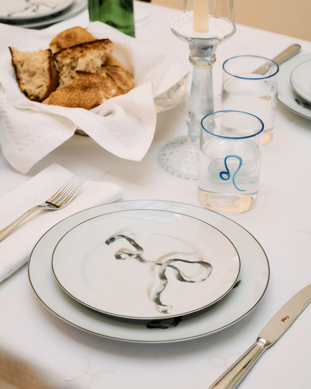 A salad plate with a black ribbon placed atop a matching dinner plate. To the left, a neatly folded white napkin lies under a silver fork, and to the right, a silver knife with a refined design. In the background, transparent hand-blown glass with cobalt blue rim and Leo astrology sign. The overall setting exudes a sense of quiet elegance.