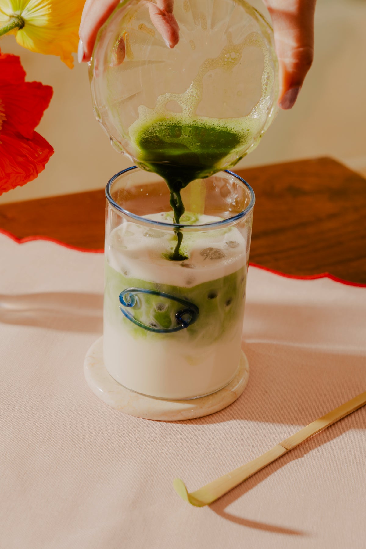 A hand is pouring a vivid matcha into a lowball glass filled with a well frothed oat milk, creating a striking green swirl as it blends. The glass has a blue Cancer zodiac symbol, and sits on a marble coaster on a wooden table with a beige cloth surface. A single lemongrass stalk lies in the foreground, leaning against the coaster. Brightly colored poppies are softly blurred in the background, adding a pop of red and yellow to the composition.