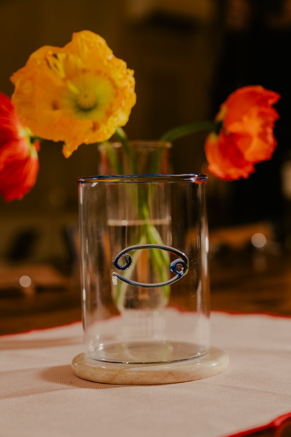 An empty clear lowball glass with a cancer zodiac sign design is centered in the foreground, placed on a marble coaster atop a beige cloth surface. The glass is framed by vibrant poppy flowers, with a yellow bloom to the left and a red one to the right, both slightly blurred, adding a lively splash of color to the scene.