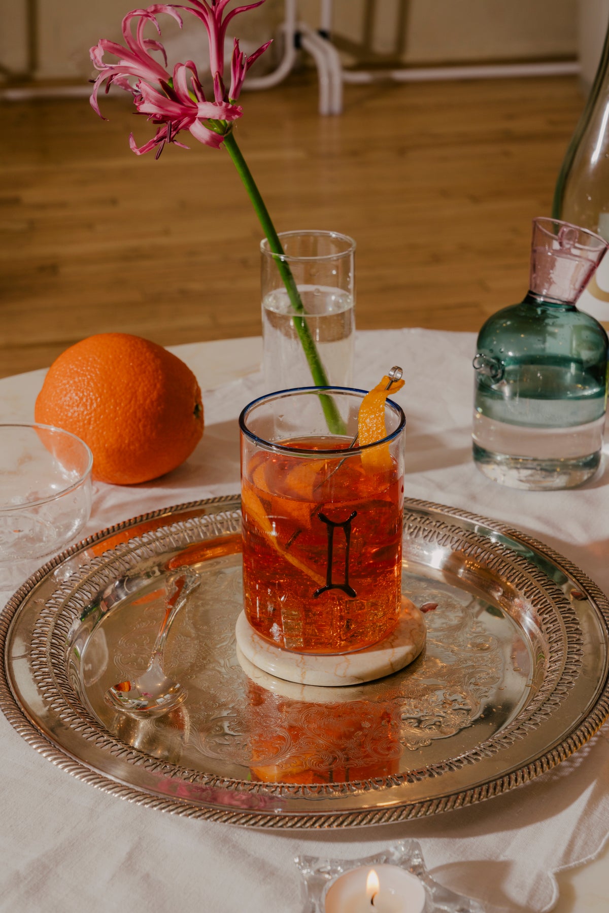 A cocktail glass adorned with a Gemini zodiac sign and rim in cobalt blue glass, with an aperol spritz, on a marble coaster atop a silver tray. An orange peel twist adds a touch of garnish. A slender vase holds a whimsical Nerine flower in hot pink. To the right, a teal oil cruet with that is filled with lemon juice for easy pouring. Beautiful colorful glassware for everyday use. Sold Individually. Murano glass tumbler is mouth-blown, hand-finished and dishwasher safe.