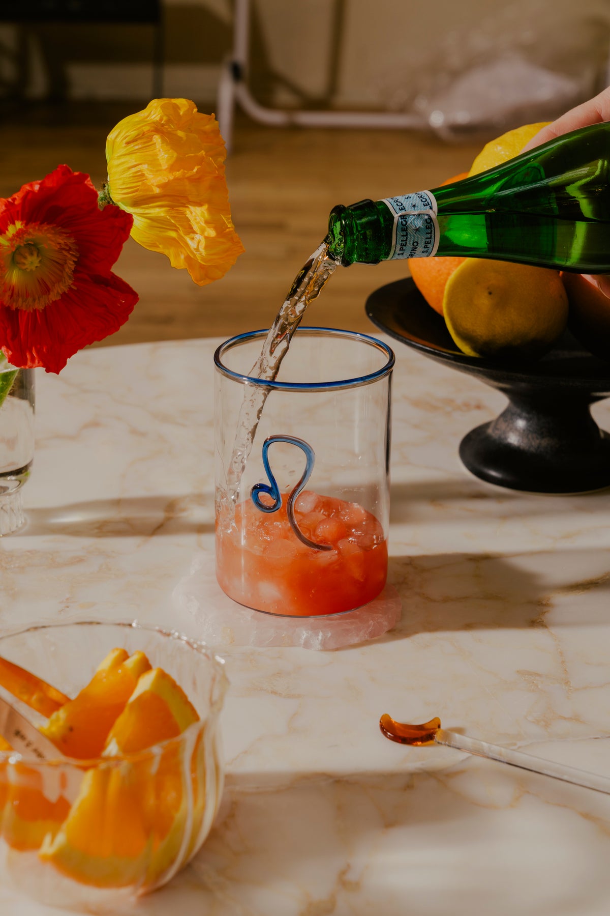 A Pomelo Margarita in a hand-blown, clear glass tumblers come with a bright twist—an almost electric lip in cobalt blue and Leo Zodiac design. In the background, a vibrant yellow poppy and a red flower add a burst of color, while a bowl of citrus fruits on a pedestal provides a backdrop for a dinner with the girlies.