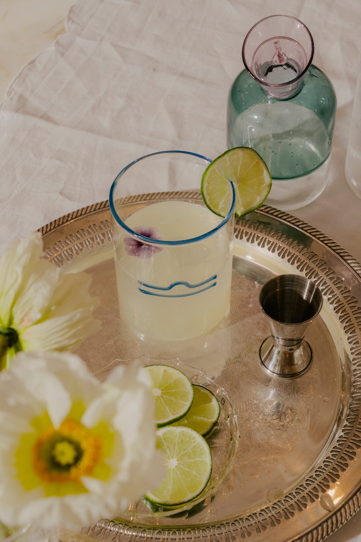 A sophisticated cocktail setup on an silver tray. A clear rocks glass with a blue rim and a libra zodiac sign has a skinny margarita, garnished with a lime wheel and a delicate purple flower floating on top. A small jigger stands to the right of the glass, ready for measuring spirits. To the left, a small dish holds several fresh lime wedges, suggesting the preparation of more drinks. Behind the main glass, a green glass bottle with a pink rim adds a contrasting color to the scene.