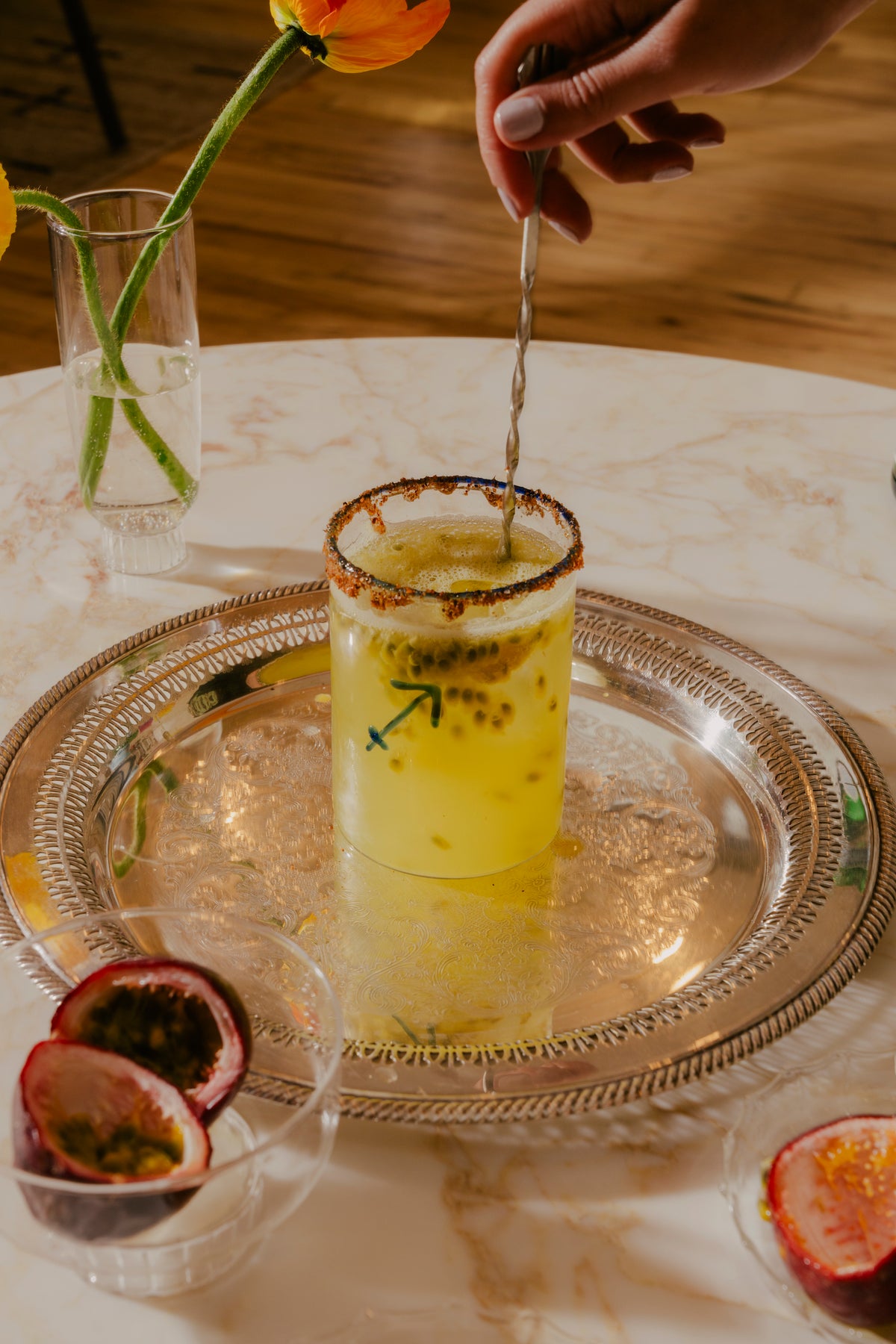 The image showcases a hand stirring a vibrant yellow cocktail adorned with a blue Sagittarius zodiac sign, presented in a clear glass with a spiced rim. The drink is speckled with passion fruit seeds, suggesting a tropical flavor profile. The glass sits atop an intricate silver tray, which reflects the light and adds to the luxurious feel of the scene. 