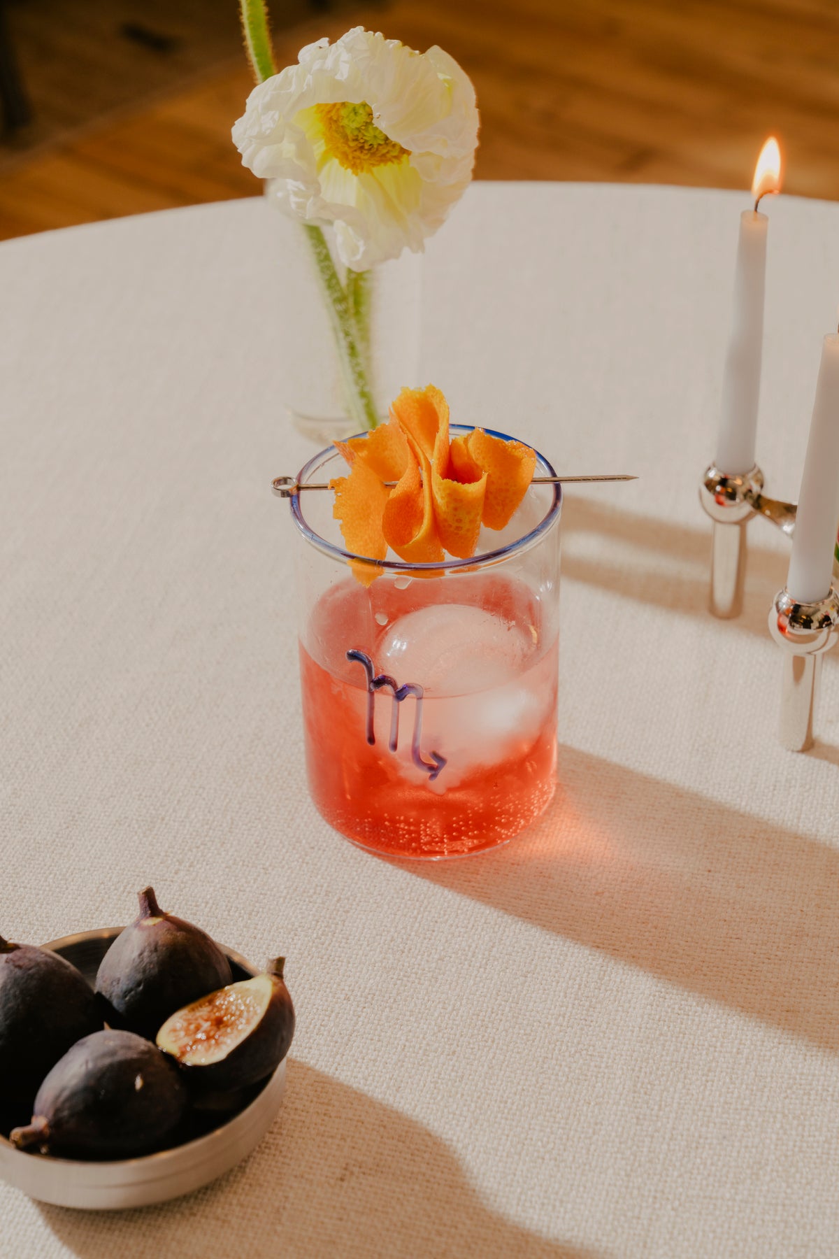 A Negroni cocktail in a clear glass with a blue rim and the astrological symbol for Scorpio in cobalt blue glass. TIn the background, a white poppy in a slender vase and a lit candle on a holder add to the serene and intimate ambiance. To the side, a bowl of ripe figs, some cut open to reveal their red interior, suggests a sophisticated and sexy ambiance for date night.