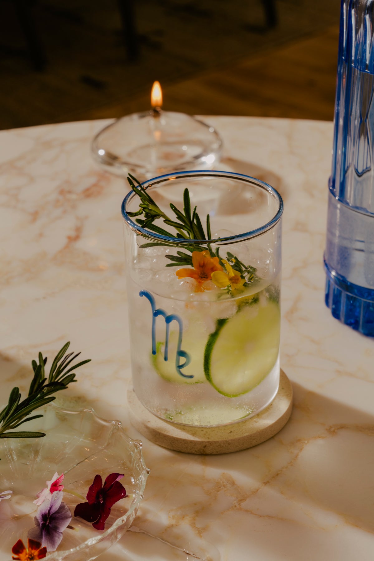 A classic and refreshing gin and tonic, served in a transparent hand-blown glass with cobalt with a blue rim and the blue astrological symbol for Virgo. The beverage is infused with green cucumber slices and a sprig of rosemary, suggesting a herbal and fresh flavor profile. A delicate edible flower floats on the surface, adding an elegant touch. These modern cocktail glasses can immediately elevate a table. Plus, since they’re so affordable, you can gift your friend a whole set of them.