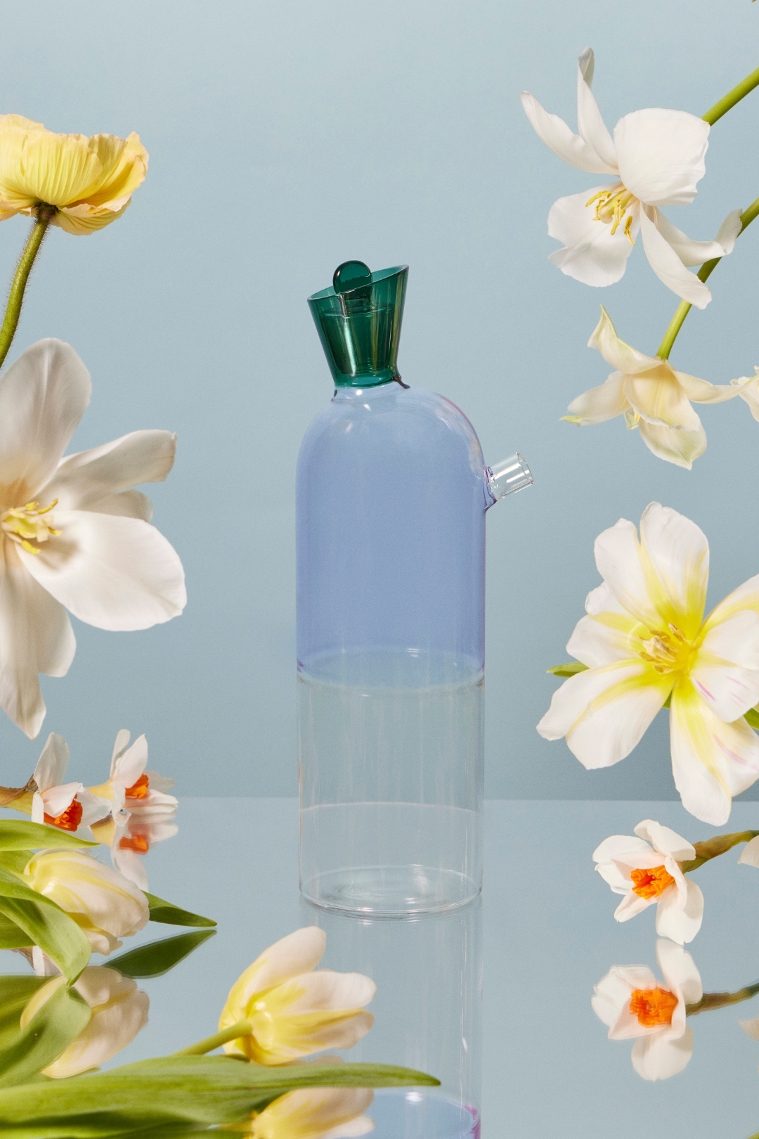 A whimsical lilac carafe and pitcher with a tapered body and anemerald green stopper, on a mirrored table. The decanter is surrounded by beautiful creamy white and soft yellow flowers, adding a touch of whimsical elegance the dinner table. 