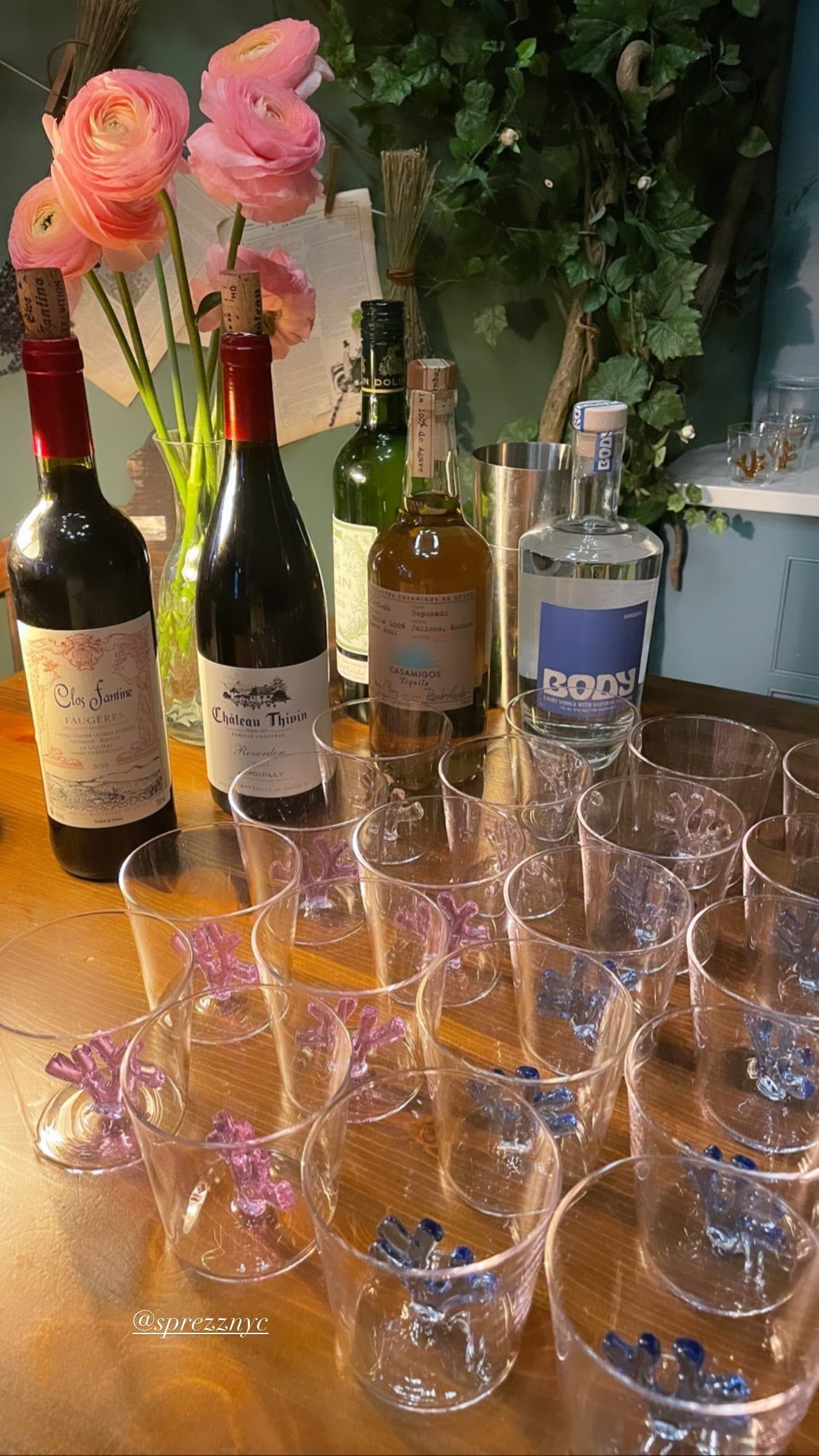 An array of Sprezz unique glassware is artfully displayed on a wooden bar, with coral sculptures in shades of lilac and blue inside of the glass. The scene is set with an eclectic mix of wine and spirits bottles, a bouquet of pink ranunculus flowers in a glass vase,  creating a vibrant and inviting atmosphere for memorable dinner parties