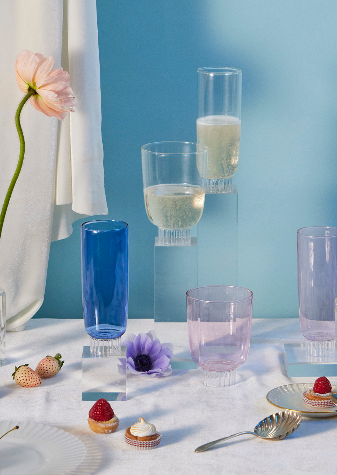 Sprezz Romantic Collection | Stemless Wine Glasses | Margherita Rui |Ichendorf Milano. Cobalt blue champagne flute, blush pink stemless wine glass, blush pink champagne flure, clear champagne glass on a table with flowers and petit fours.