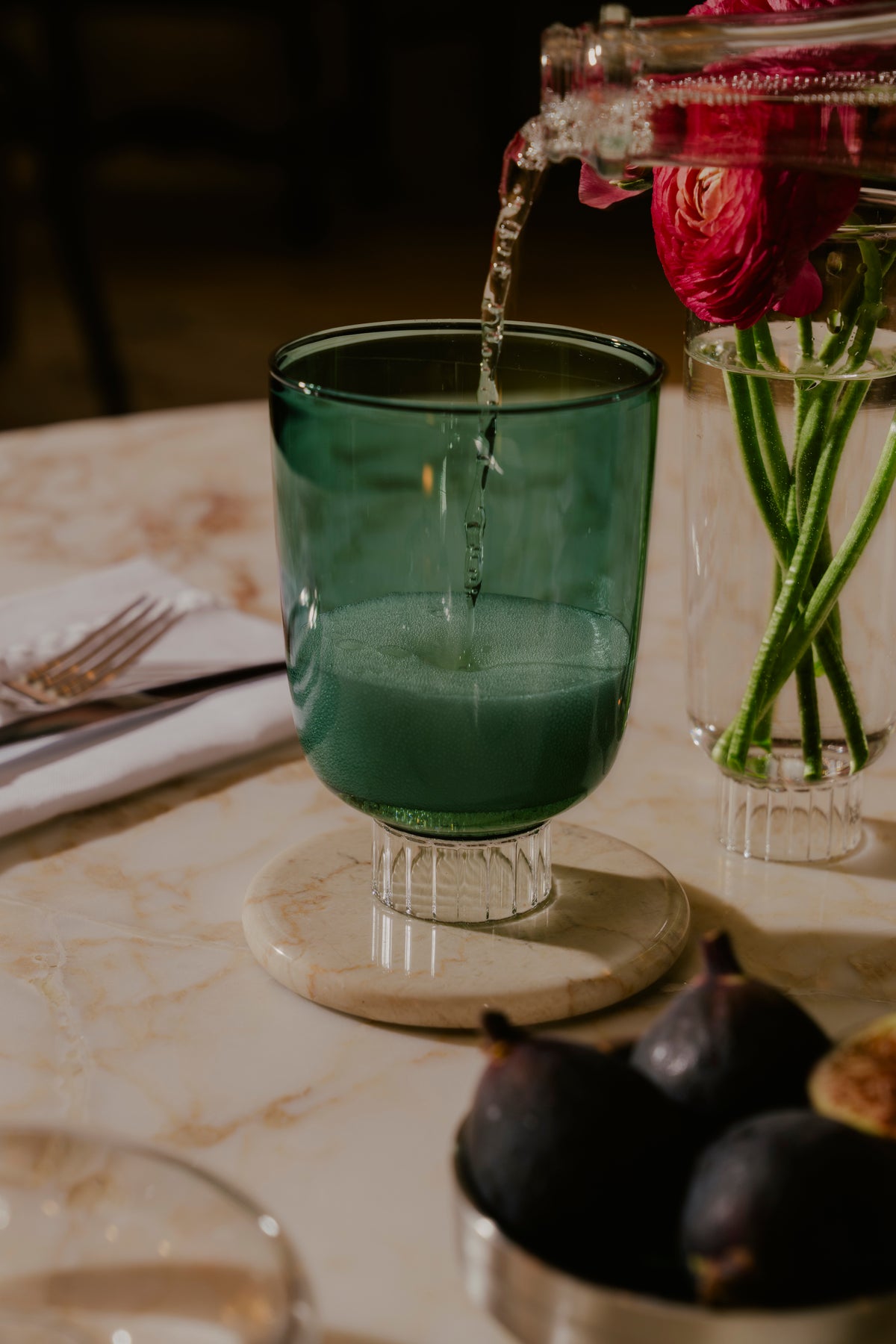An emerald green stemless wine glass with a ribbed base, set on a marble table. The glass, part of a sophisticated table setting, is accompanied by a vase of pink ranunculus flowers and figs, capturing the essence of elegant dining.