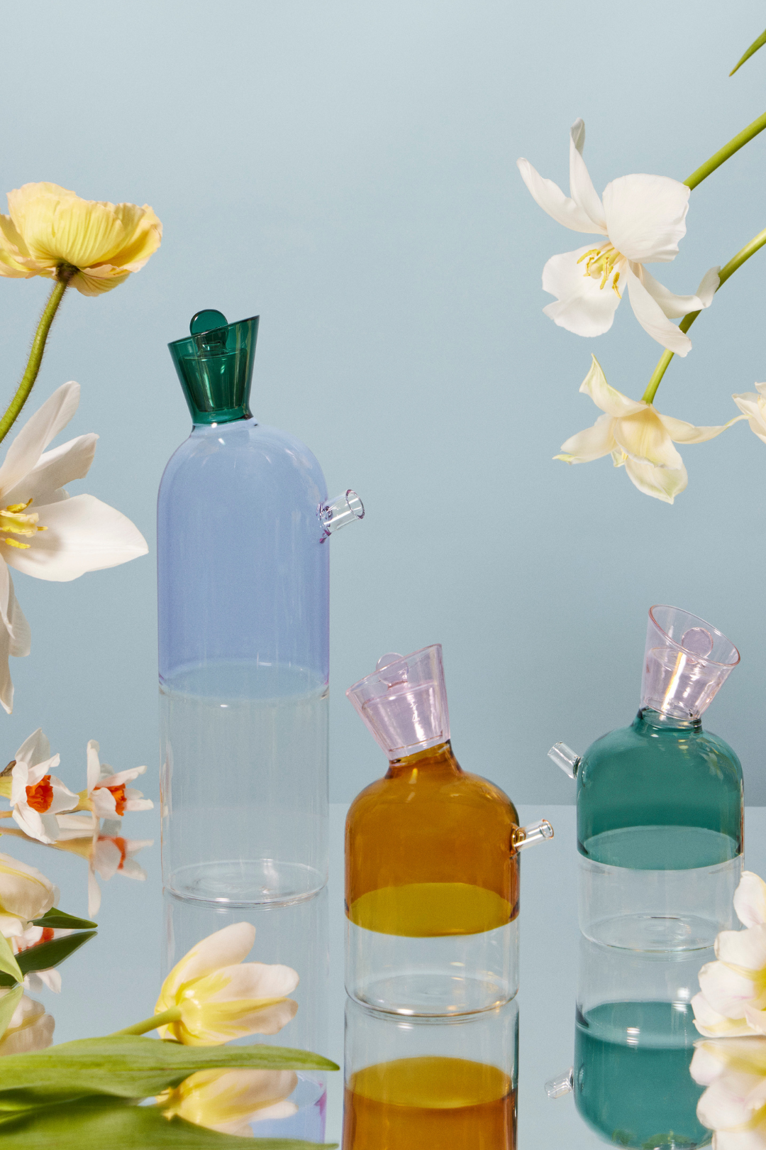 Lilac Carafe and two oil and vinegar bottles in teal and amber borosilicate glass on a mirrored table surrounded by tulips and daffodils. Contemporary, minimalist allure animates the modernist design of these colored glass decanters